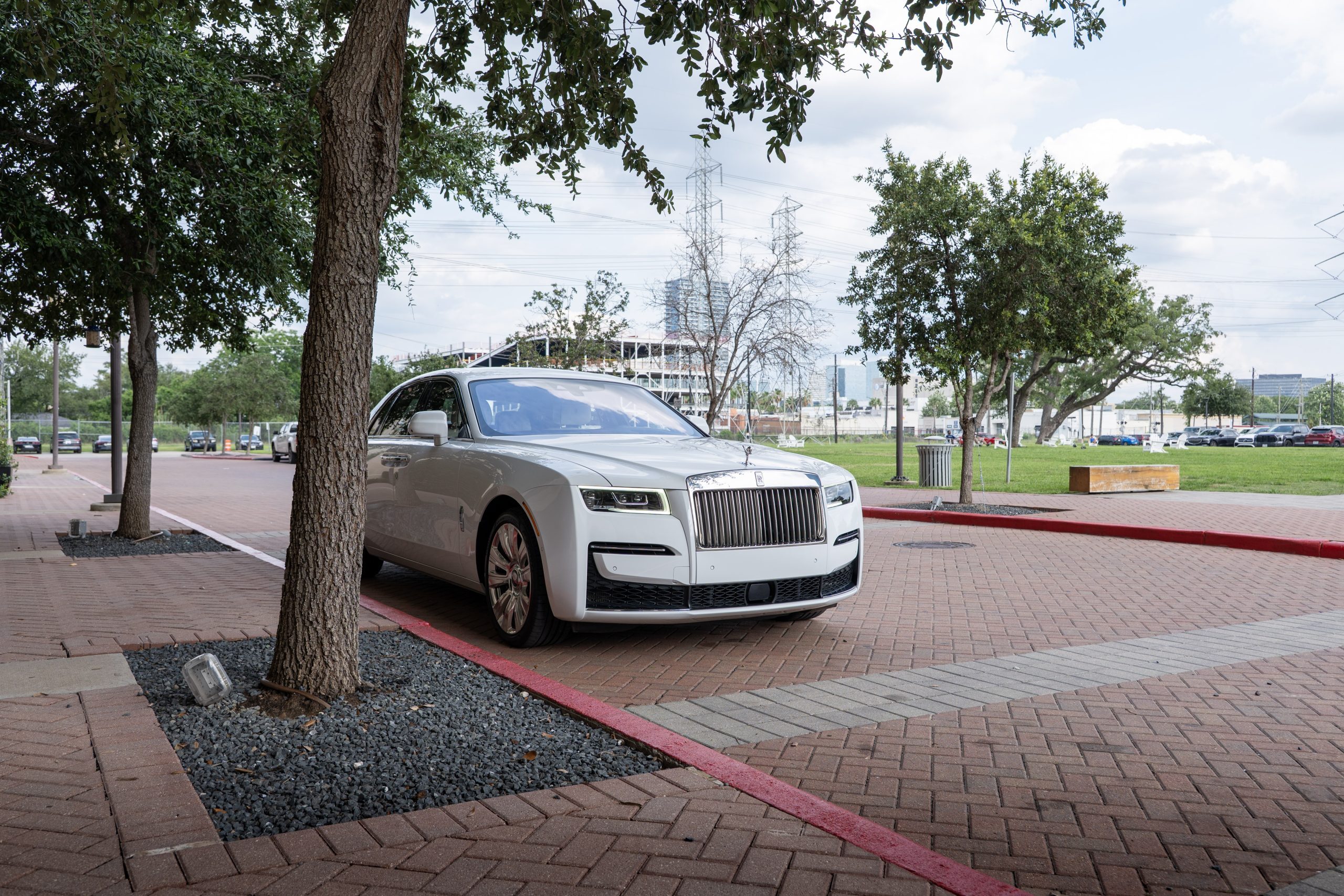 White Rolls Royce Ghost parked on city street, partial tree cover and modern Houston skyline in the background.