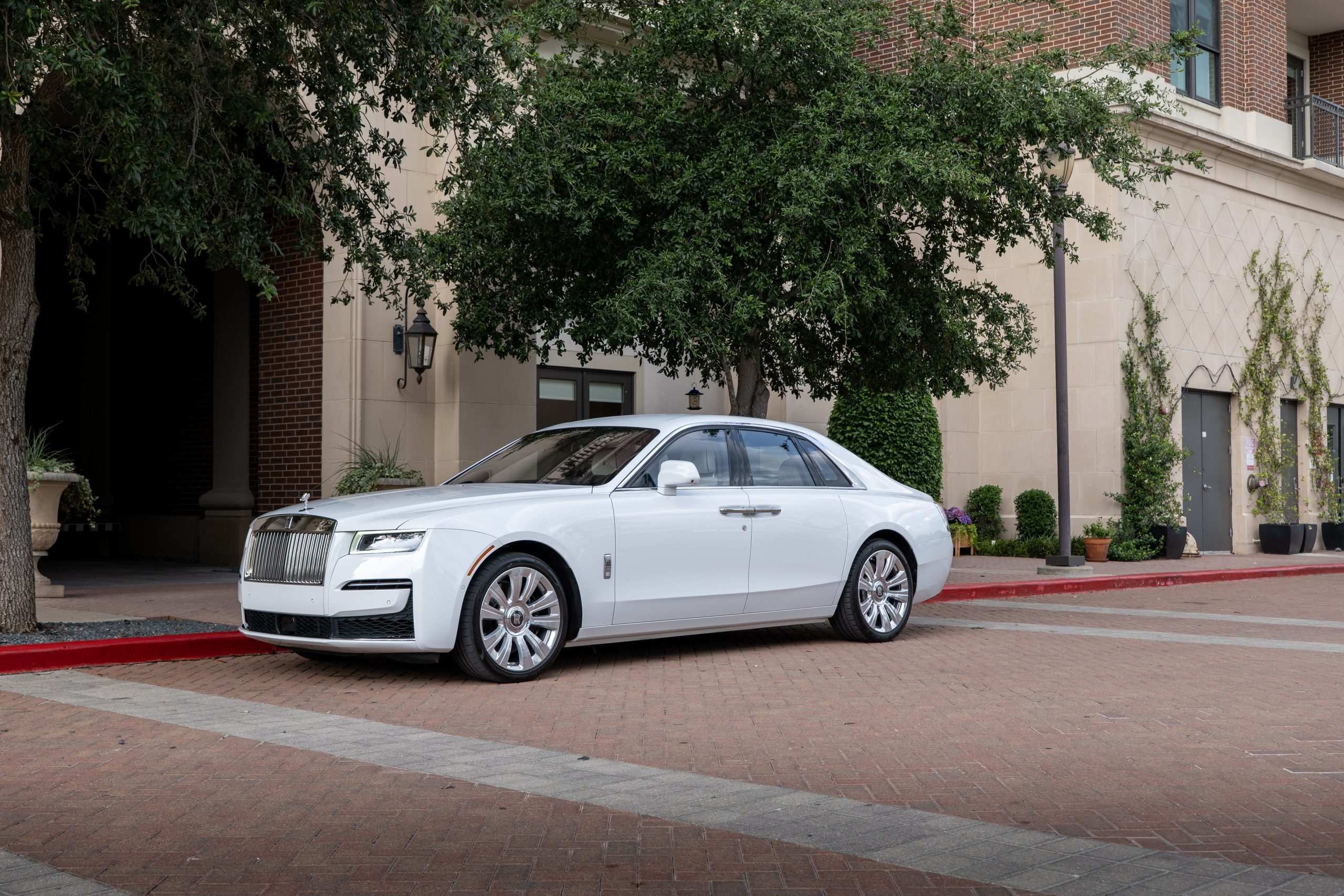 Full side view of white Rolls Royce Ghost sedan showing off classic silhouette, luxury wheels, and urban backdrop.