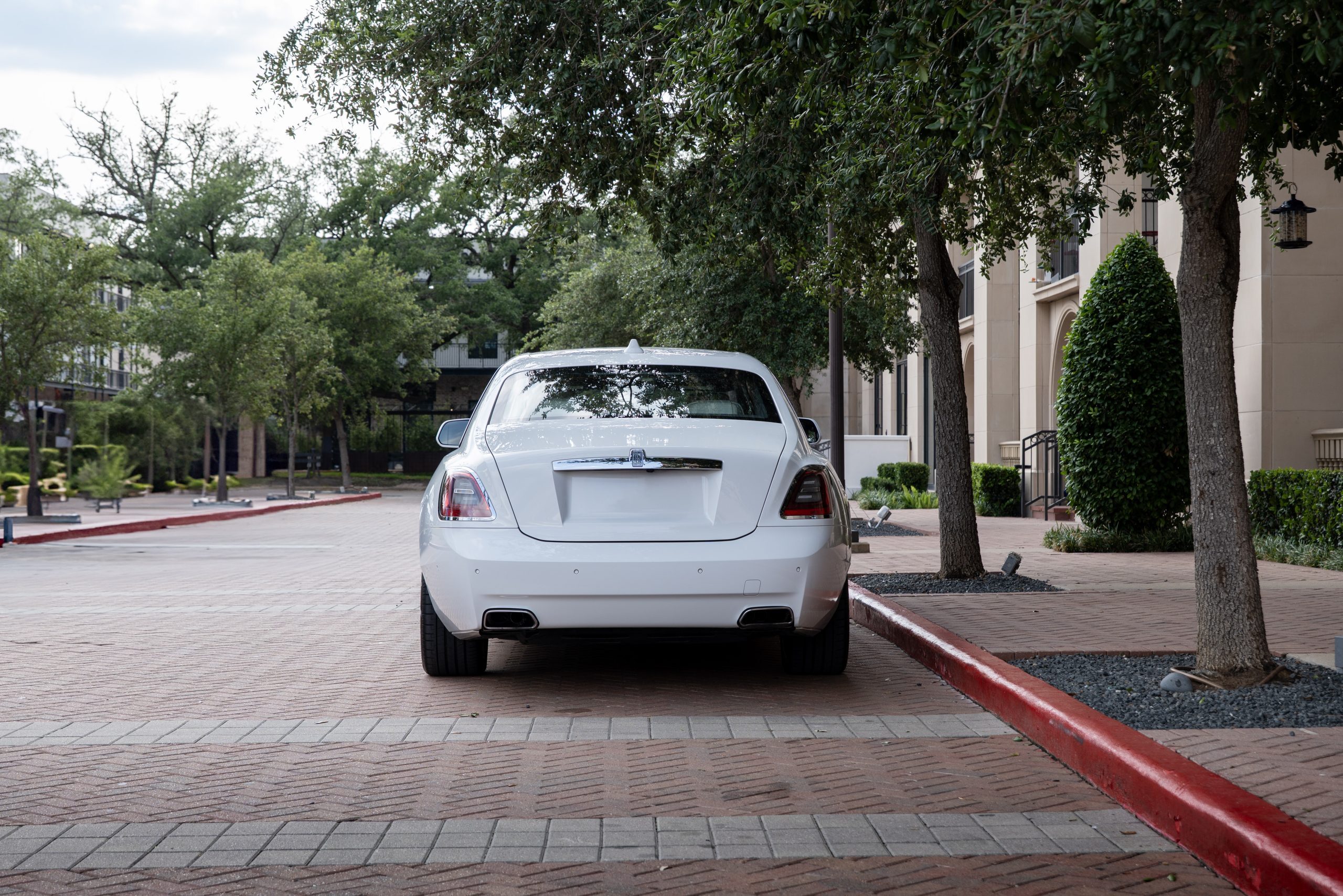 Rear three-quarter view of white Rolls Royce Ghost parked near luxury residences.