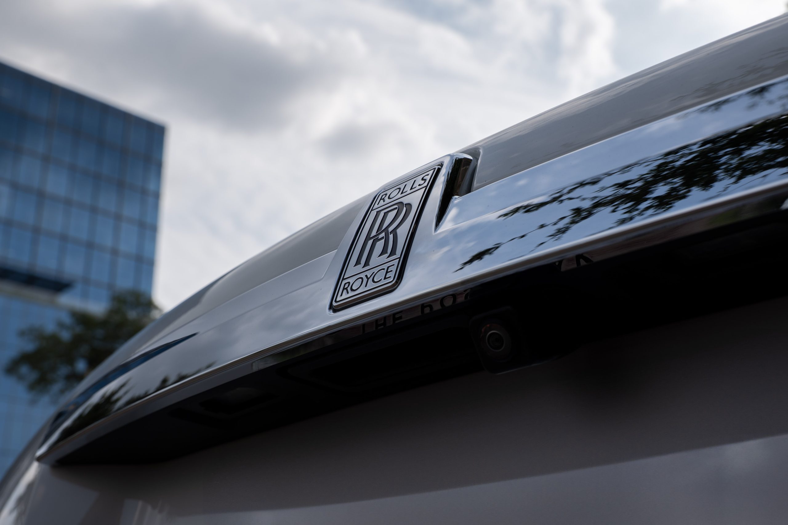Polished chrome Rolls Royce badge on trunk, close-up showing urban skyline reflections and reverse camera.