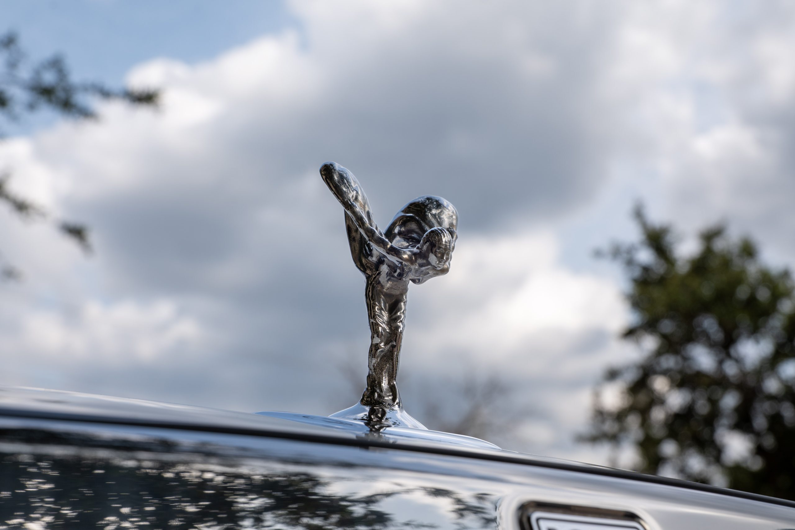 Close-up of Spirit of Ecstasy hood ornament on Rolls Royce Ghost, with blurred clouds and trees in background.