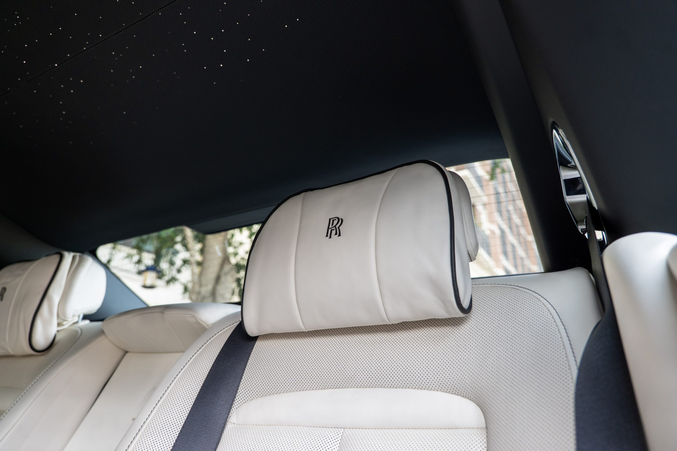 Close-up of Rolls Royce Ghost rear seat white leather headrest with RR embroidery, set beneath starlight headliner.