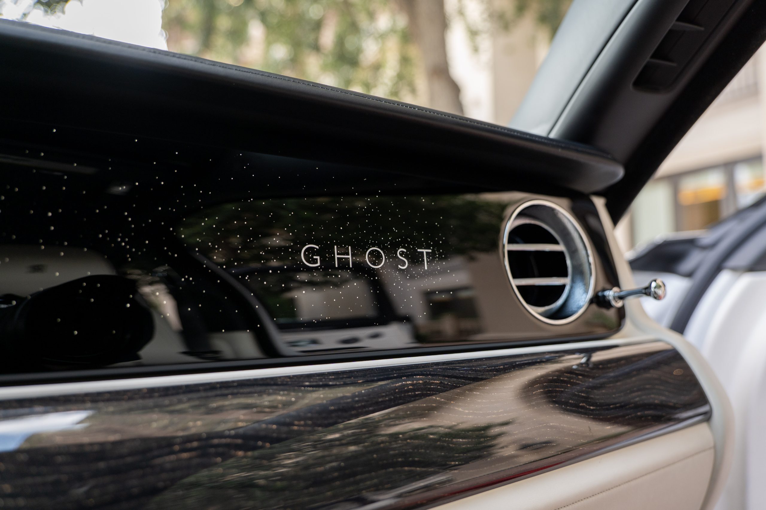 Rolls Royce Ghost dashboard with illuminated "GHOST" logo and starlight effect beside chrome vent and wood trim.