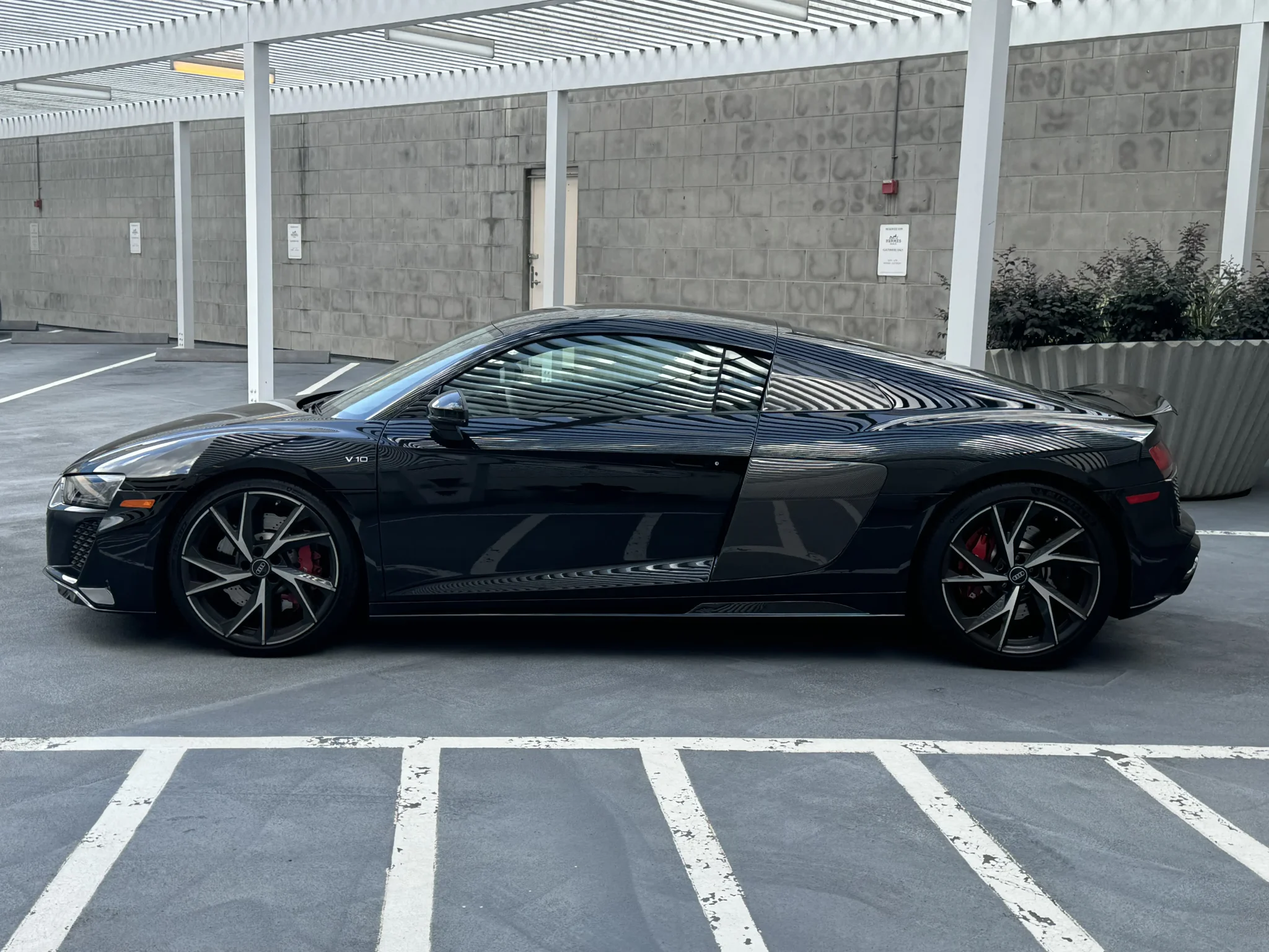 Side profile of a black Audi R8 parked in a covered, urban parking lot, showcasing its low-slung stance, angular body lines, and distinct alloy wheels.