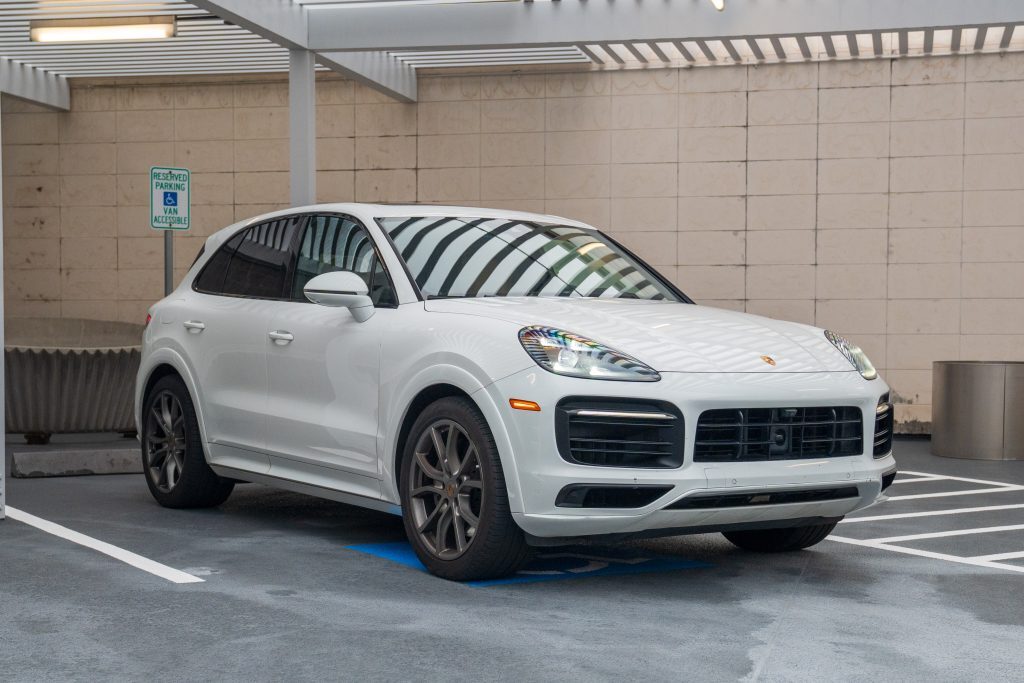 A white Porsche Cayenne S is photographed from the front three-quarter angle, parked in a reserved accessible space under a modern canopy with striped shadows.